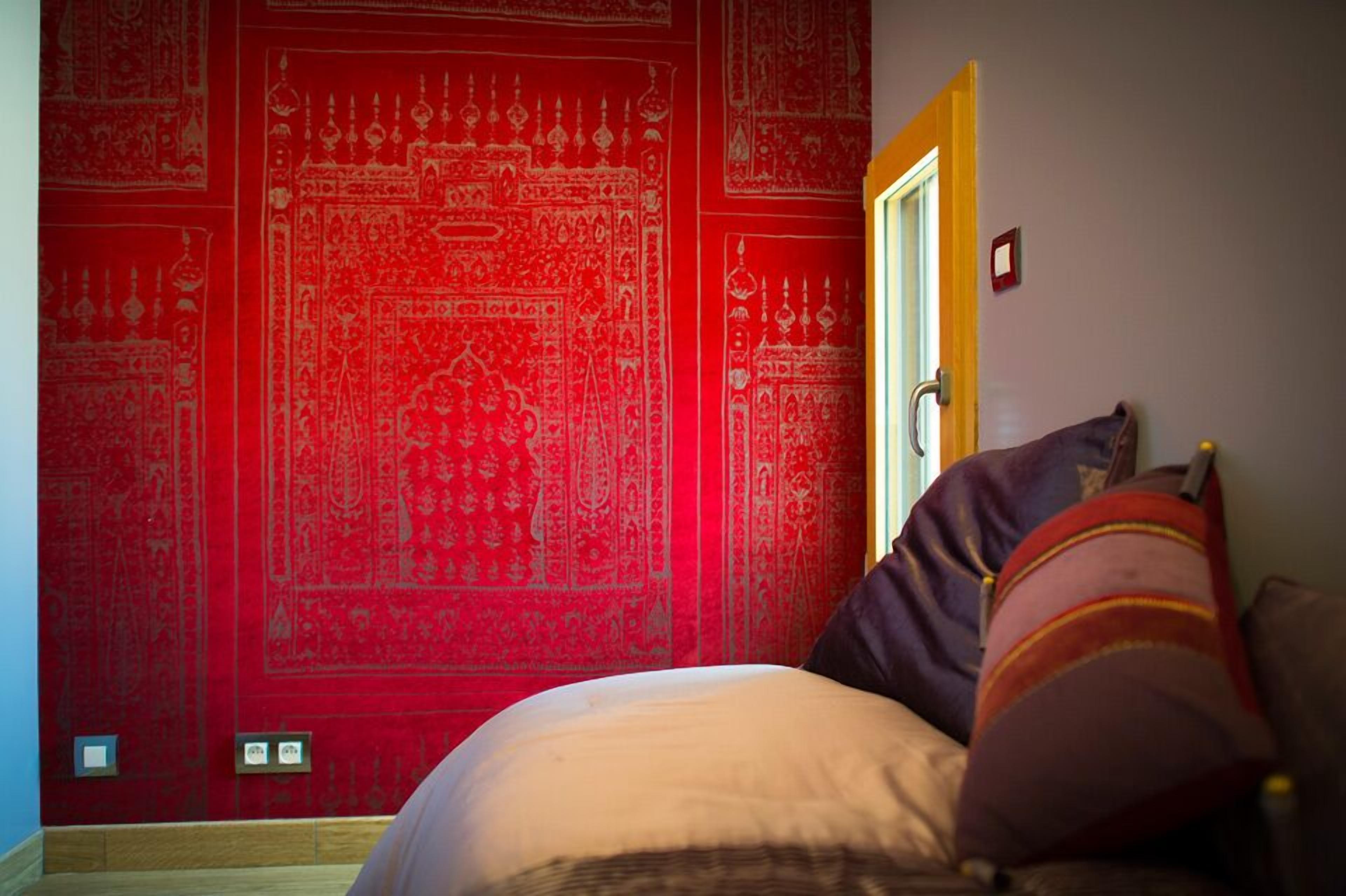 Een kamer met een rode, behang met patroon, een bed met kussens en een deur.