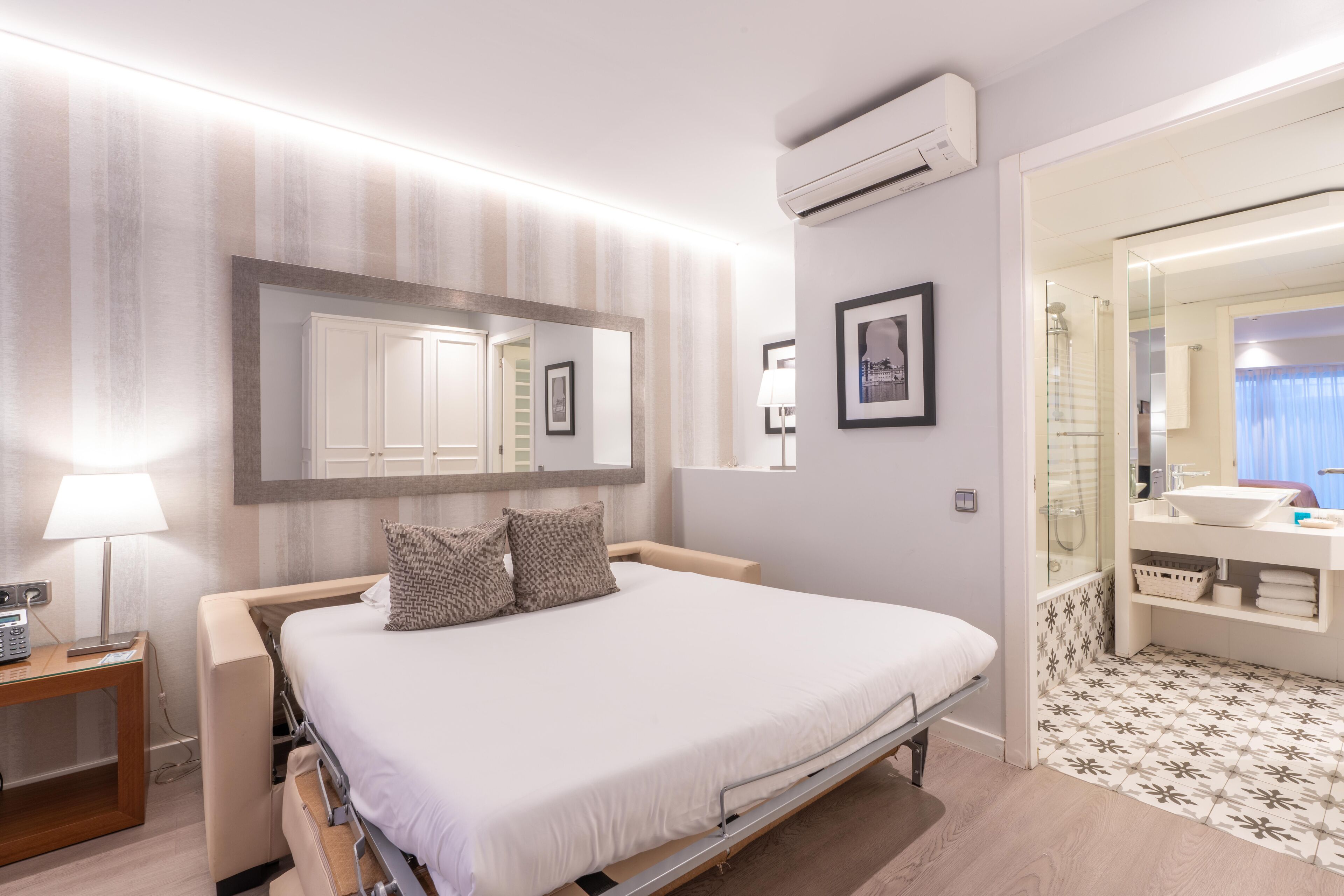 A hotel room with a bed, bedside table, lamp, air conditioning unit, and a bathroom with a mirror, sink, and patterned floor.