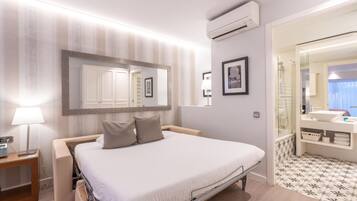 A hotel room with a bed, bedside table, lamp, air conditioning unit, and a bathroom with a mirror, sink, and patterned floor.