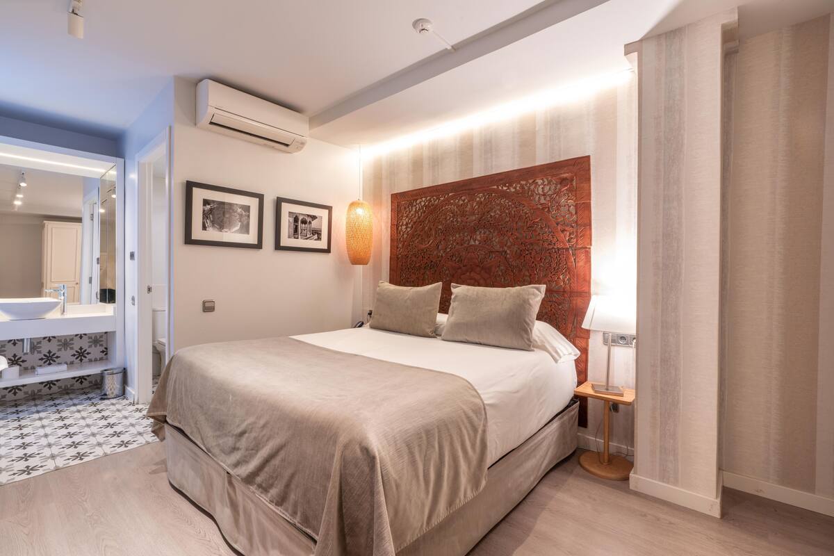 A modern bedroom with a large bed, a bedside table, a wall-mounted air conditioner, and a bathroom with a sink and mirror.
