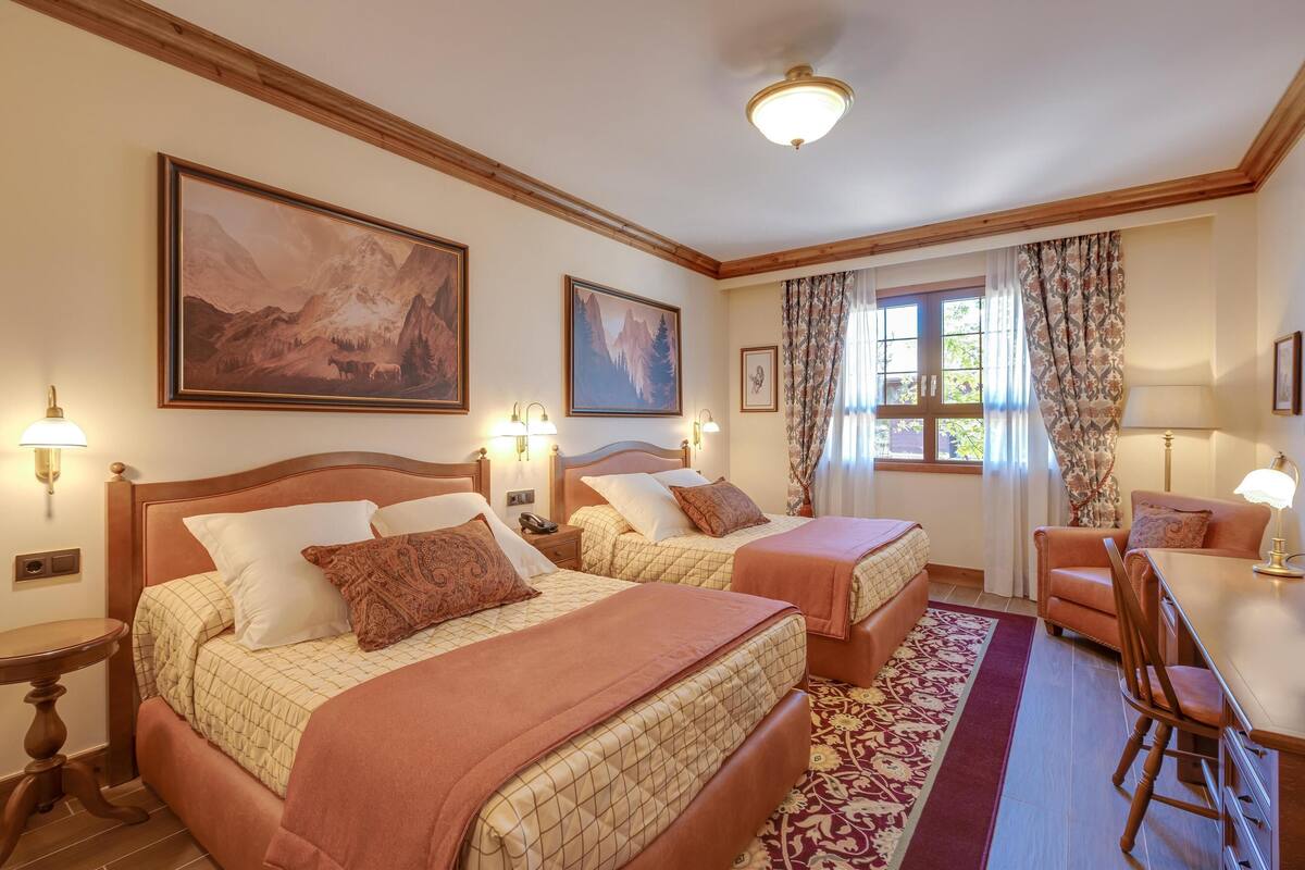 Une chambre d’hôtel avec deux lits, un bureau et une chaise. Les murs sont ornés de tableaux, il y a une fenêtre avec des rideaux et le plafond est en bois.