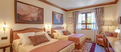 A hotel room with two beds, a desk, and a chair. There are paintings on the walls, a window with curtains, and a wooden ceiling.