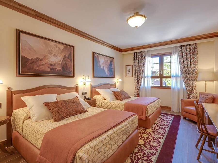 Habitación de hotel con dos camas, un escritorio y una silla. Hay cuadros en las paredes, una ventana con cortinas y techo de madera.