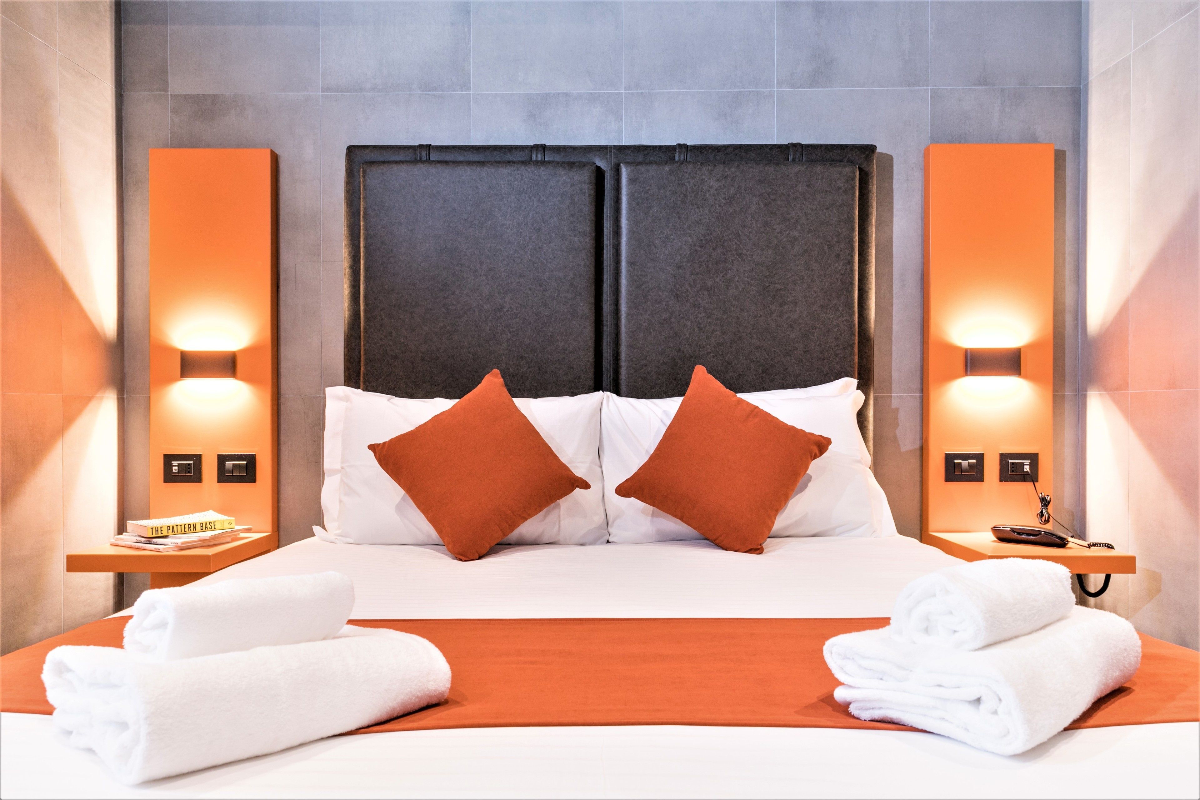 A hotel room with a large bed, two pillows, a nightstand with a book, a phone, and a towel.