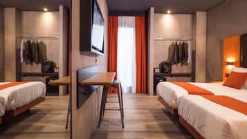 Une chambre d’hôtel avec deux lits, un bureau et une chaise. On y trouve deux tables de chevet avec des lampes, un téléviseur fixé au mur et une fenêtre avec des rideaux.