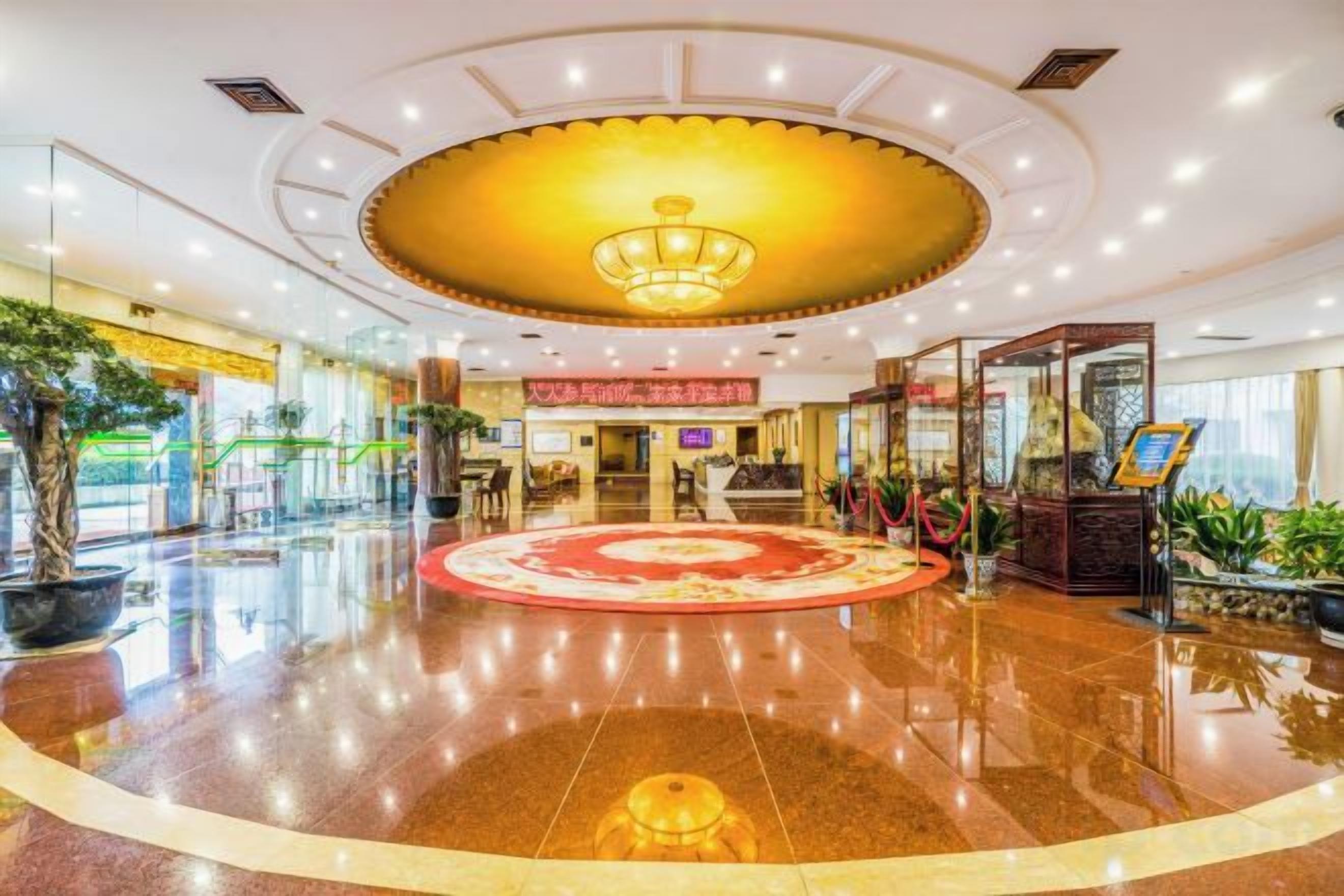 Un hall de hotel con una alfombra circular en el centro, mostrador de recepción y plantas en macetas.