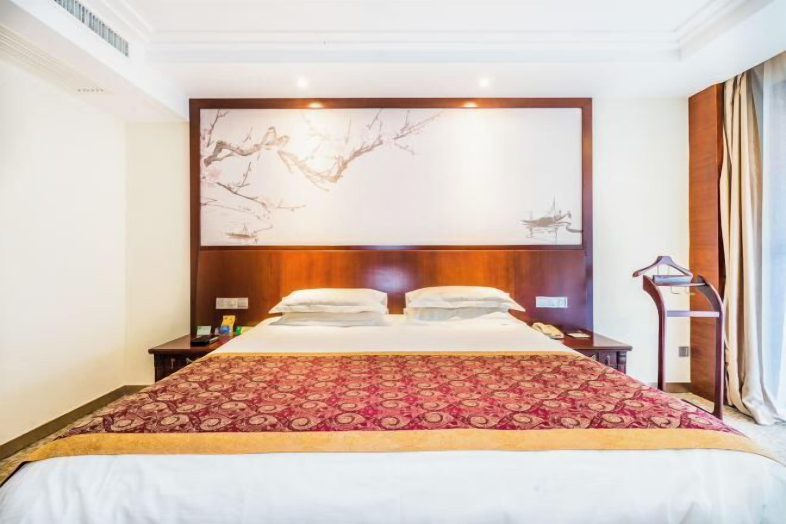 A hotel room with a large bed, a wooden headboard, a wall-mounted artwork, and a bedside table with a lamp.