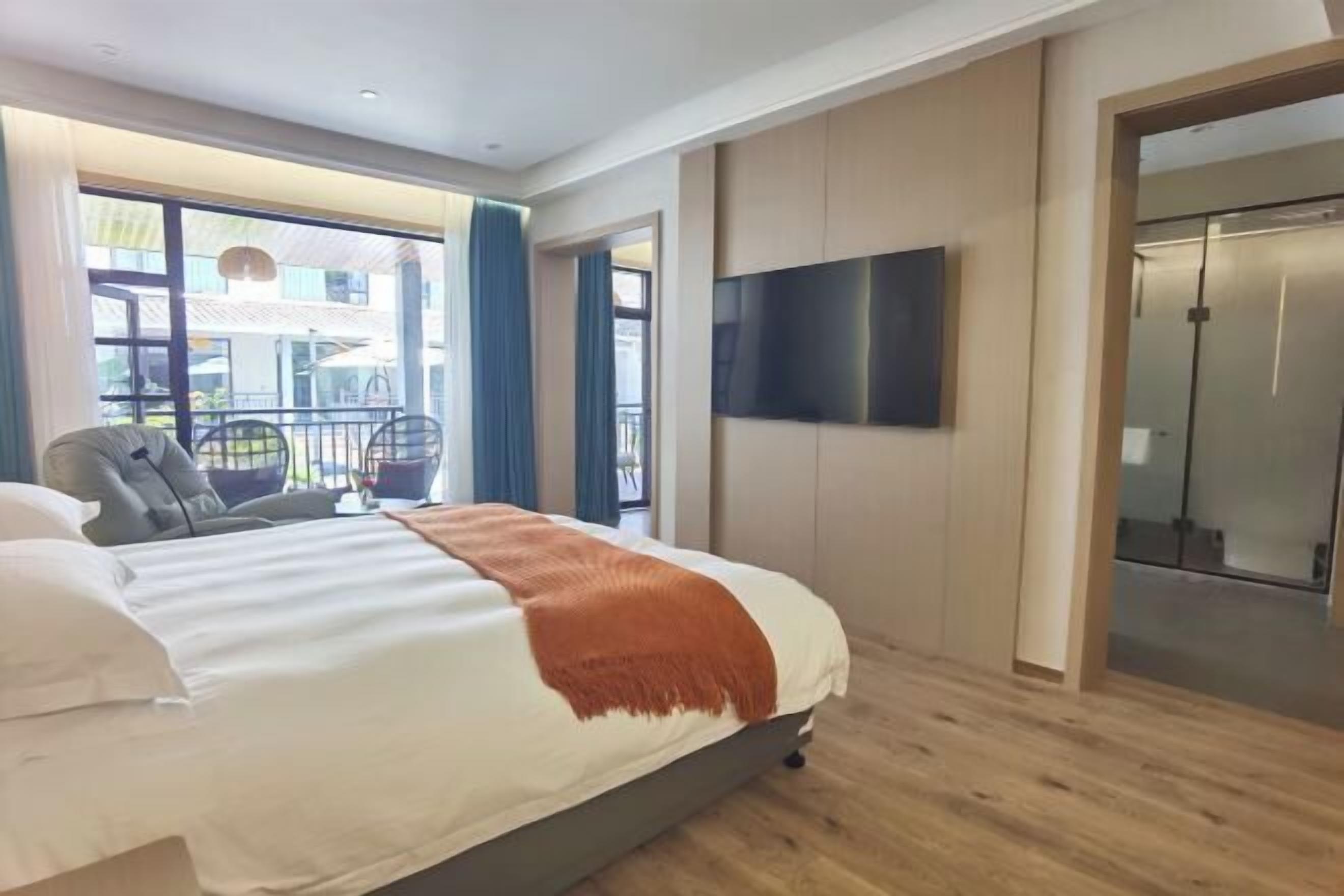 A hotel room with a bed, a TV mounted on the wall, a balcony with a chair, and a sliding door.