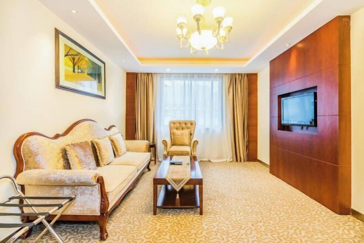 A hotel room with a sofa, armchair, coffee table, TV mounted on a wooden wall, and a painting on the wall.