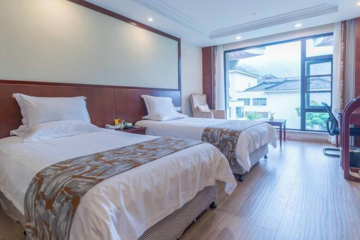 A hotel room with two beds, a chair, a table, and a large window with a view.