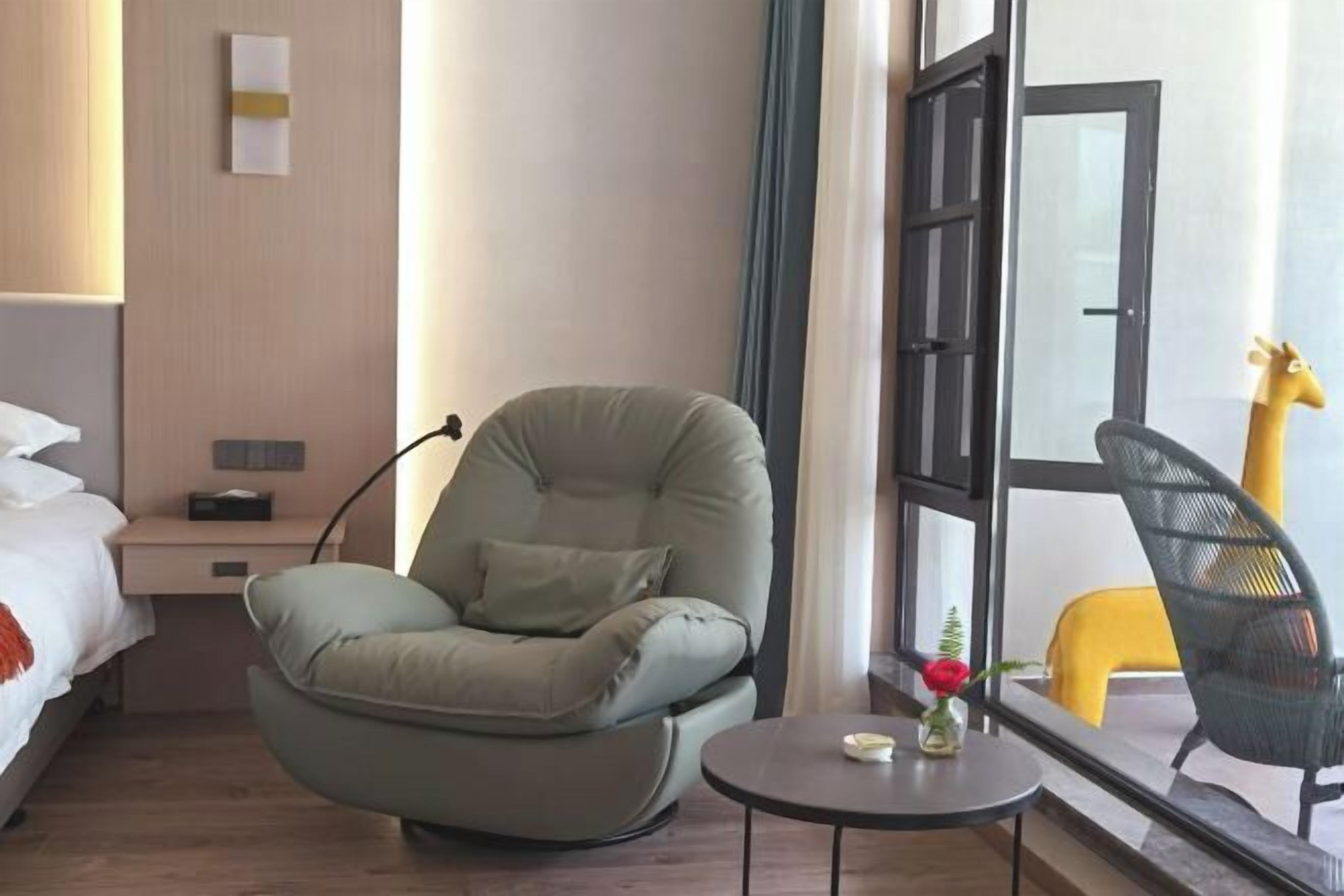 Una habitación de hotel con una cama, un sillón verde, una mesita redonda con un florero y una ventana que ofrece vistas a una estatua de jirafa.