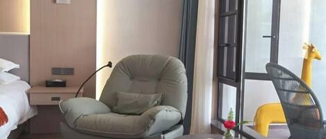 Una habitación de hotel con una cama, un sillón verde, una mesita redonda con un florero y una ventana que ofrece vistas a una estatua de jirafa.