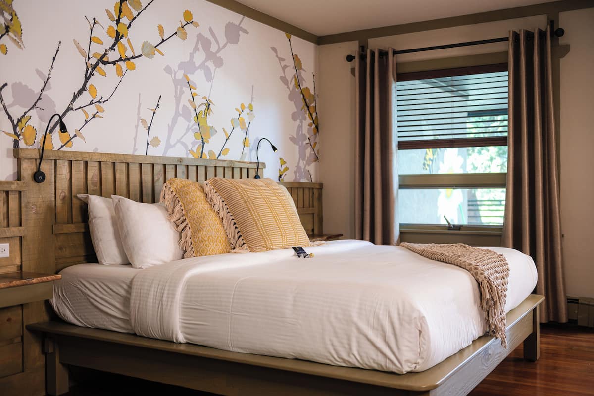 A neatly made bed with white linens and a beige throw blanket, a wooden headboard, and a wall mural of branches.