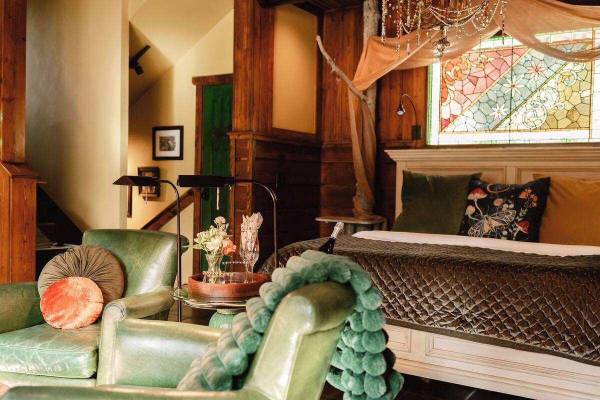 A room with a green leather armchair, a bed with a quilted headboard, a chandelier, and a stained glass window.