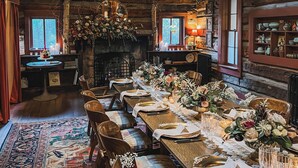 A rustic dining room with wooden beams, a long table set for a meal, and a fireplace with a flower arrangement. - Highland Haven Creekside Inn (Evergreen)