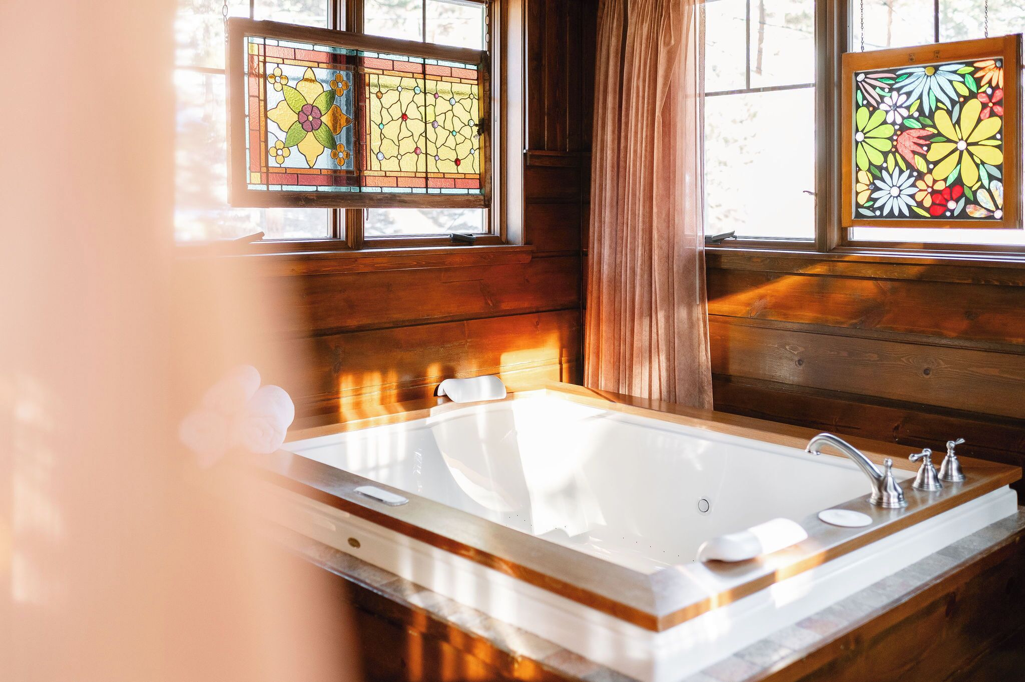 Interior de una cabaña de madera con una bañera grande, dos ventanas de vidrio emplomado y una pared de madera.