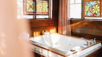 Interior de una cabaña de madera con una bañera grande, dos ventanas de vidrio emplomado y una pared de madera.