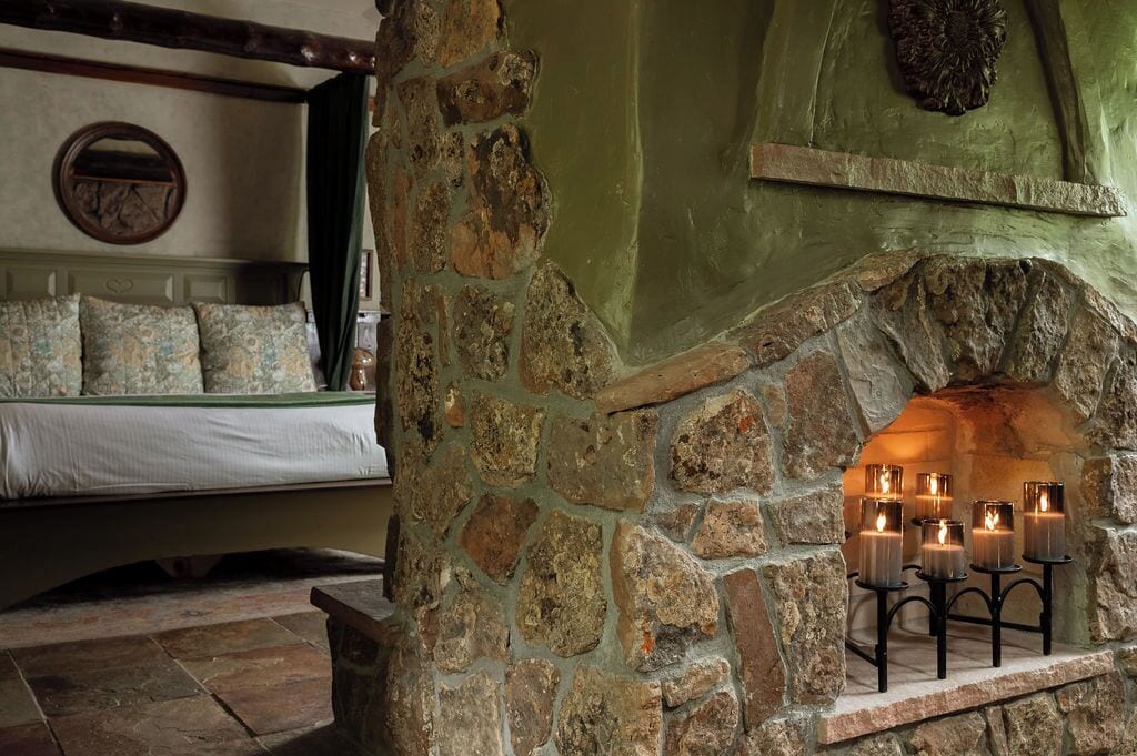 Una chimenea de piedra con velas, una cama con almohadones estampados y decoración rústica en la pared.