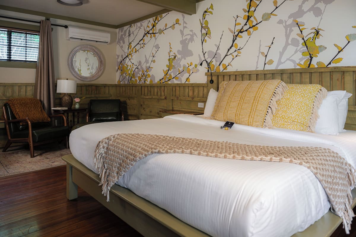 A neatly made bed with a patterned throw, a wooden headboard, and a wall with a floral mural.