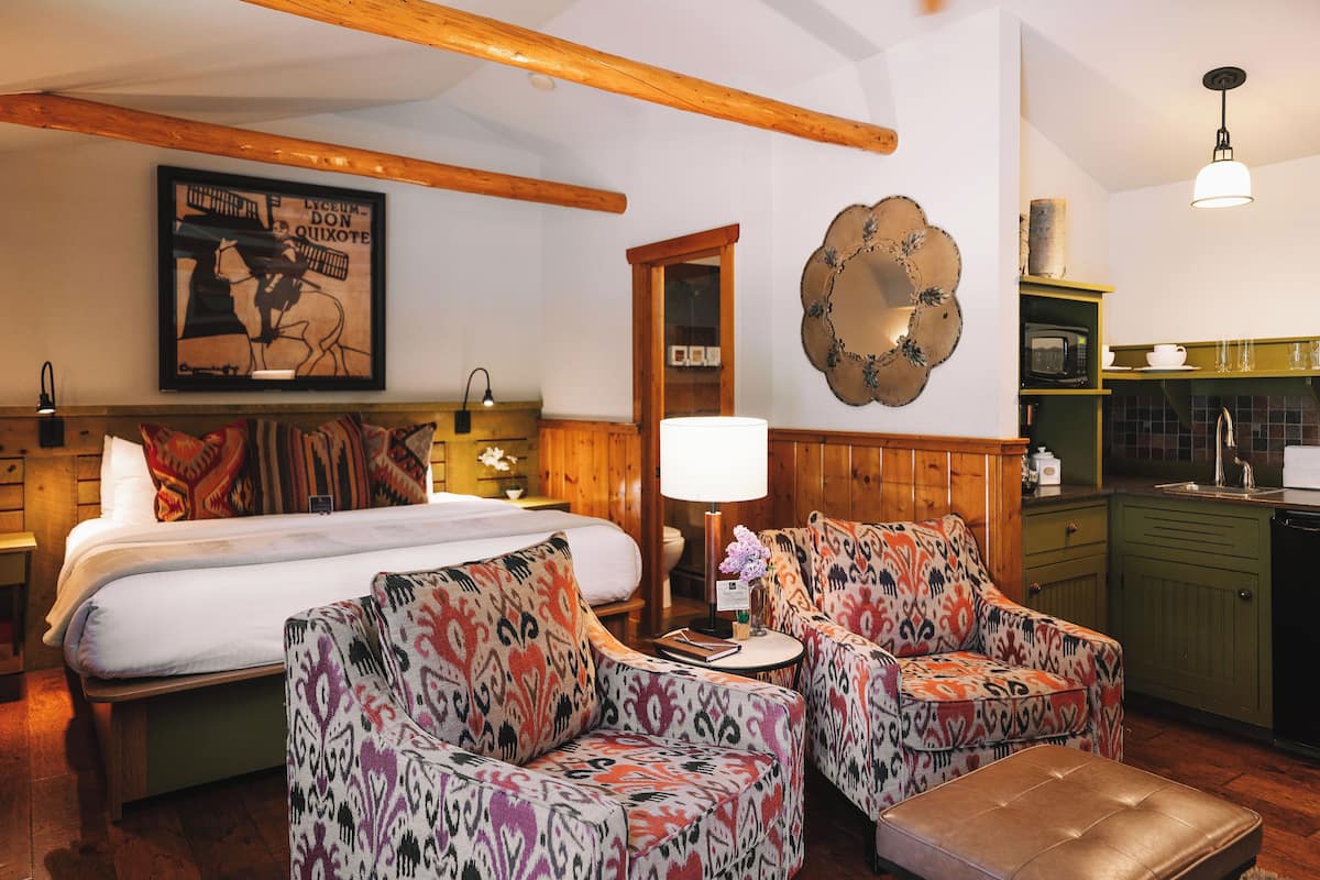 A room with a bed, two patterned armchairs, a small table with a lamp, a mirror, and a kitchen area with a microwave.