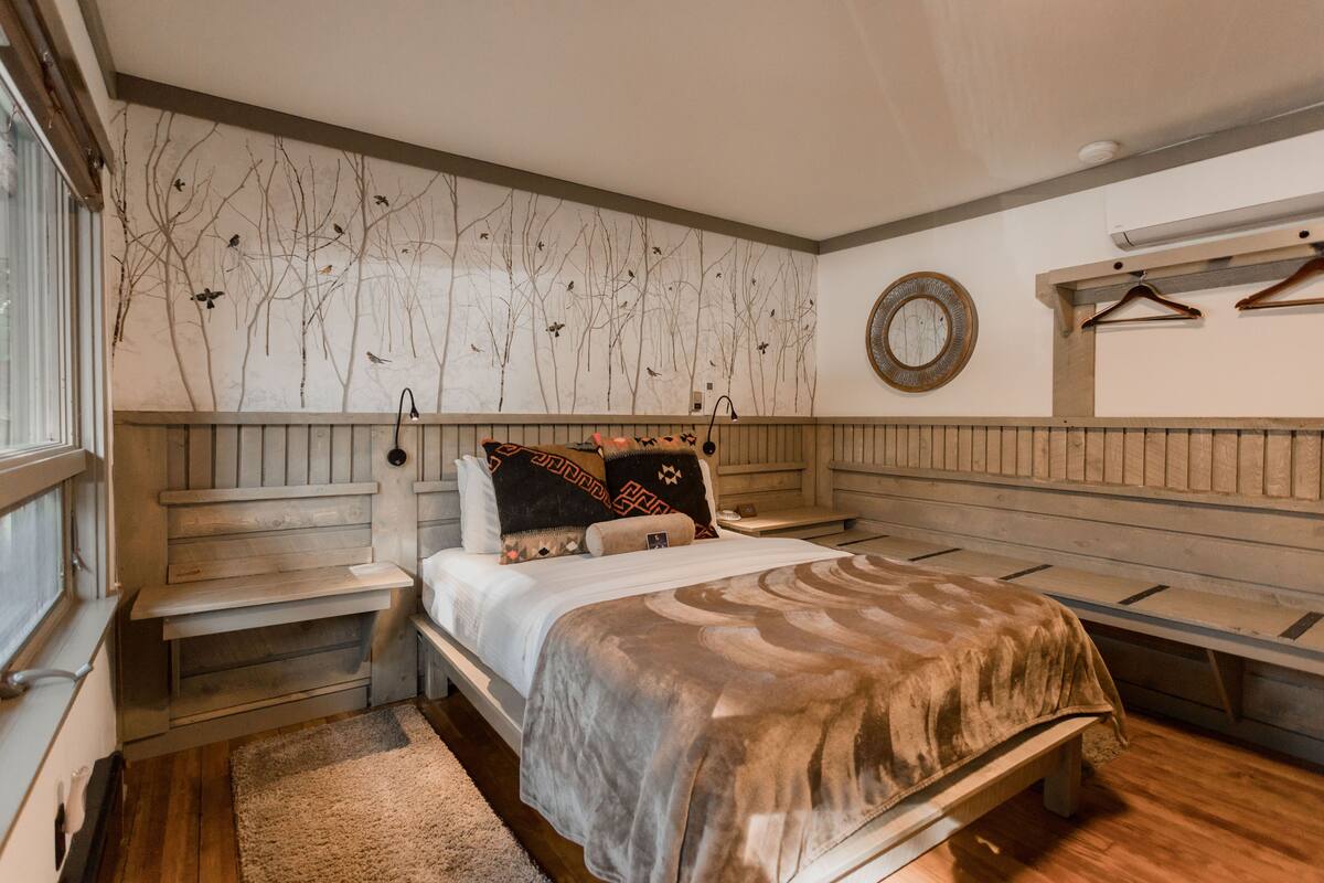 A bedroom with a bed, a nightstand, a wooden bench, a wall-mounted mirror, and a window.