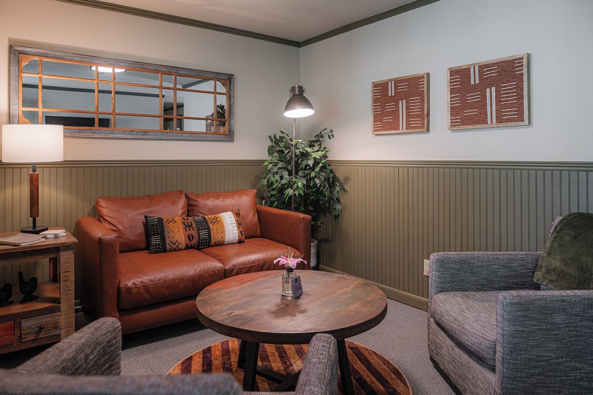 A living room with a brown leather sofa, a round wooden coffee table, and a gray armchair. A floor lamp and a potted plant are also visible.