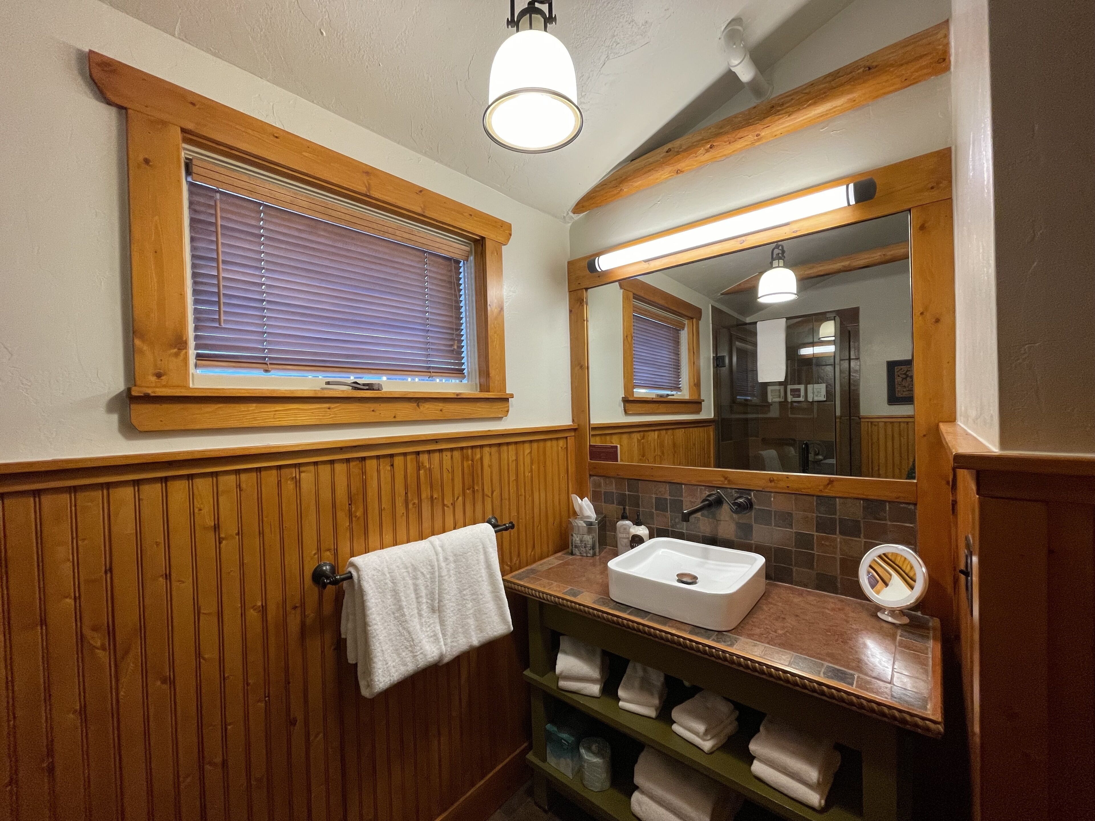 Un baño con paneles de madera, una ventana con persianas, un lavamanos con espejo y un perchero para toallas.