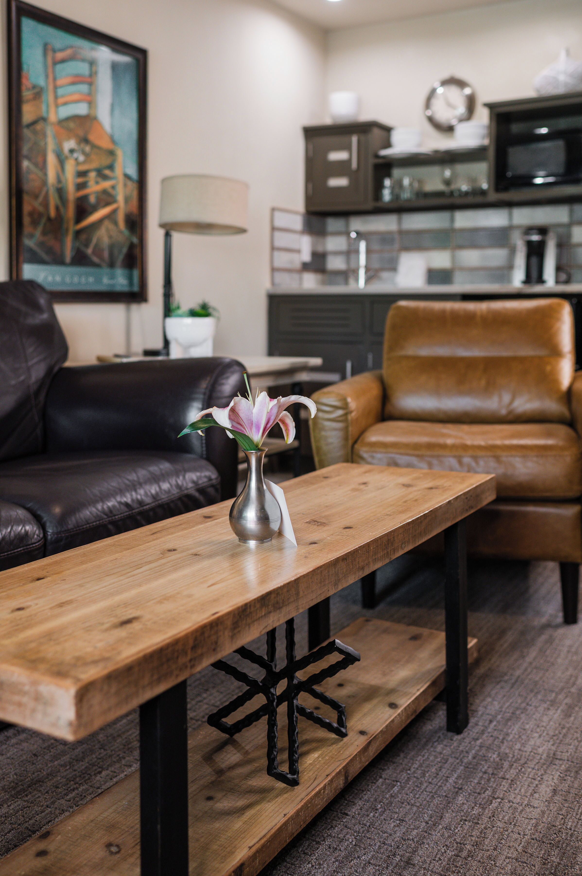 A living room with a wooden coffee table, a vase with flowers, a brown leather sofa, and a kitchen area in the background.