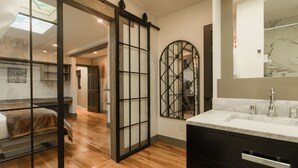 A modern bathroom with a large mirror, wooden flooring, and a sliding glass door leading to another room. - Highland Haven Creekside Inn (Evergreen)