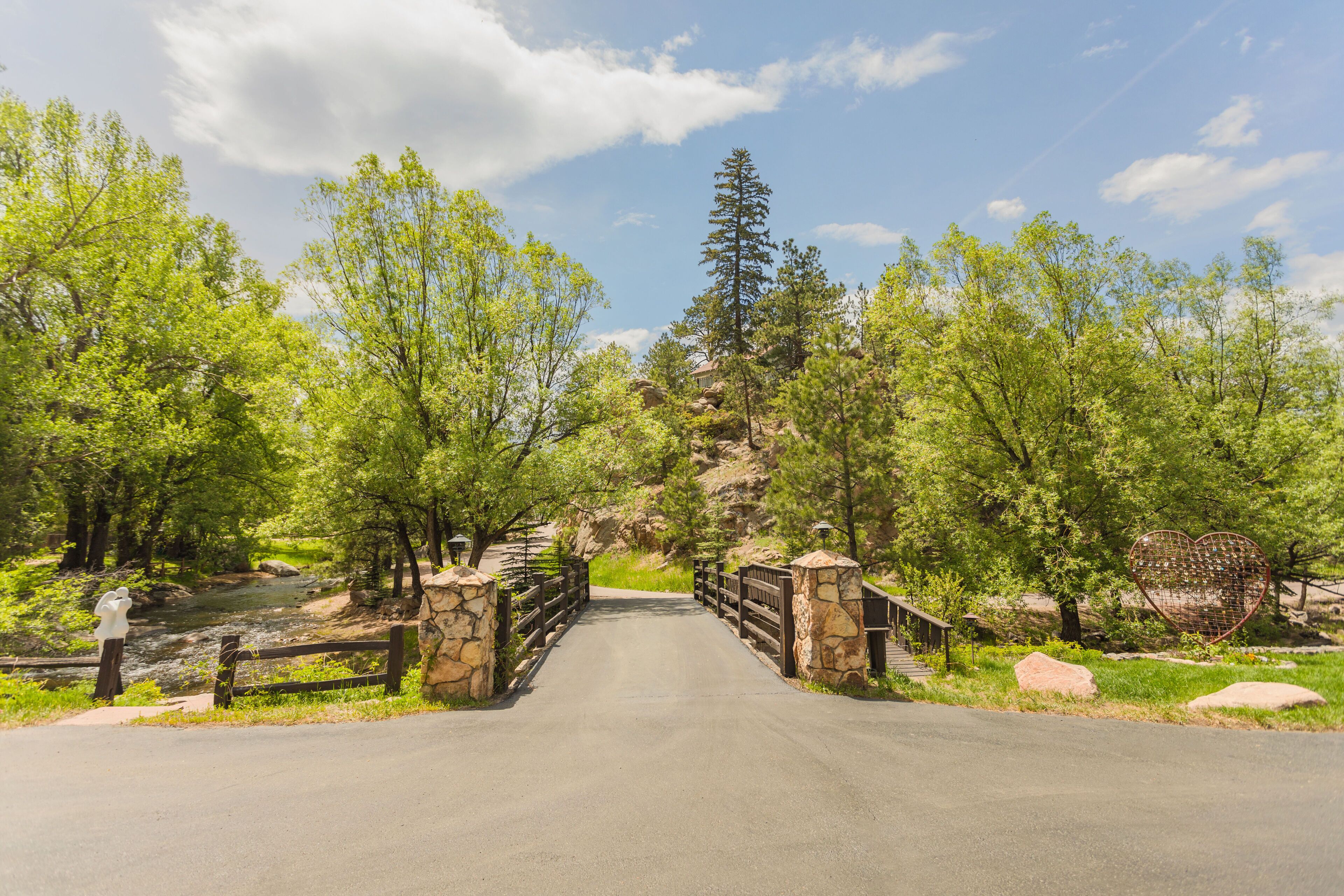 A paved road leading to a stone bridge with a metal railing, surrounded by green trees and a rocky hill in the background.
