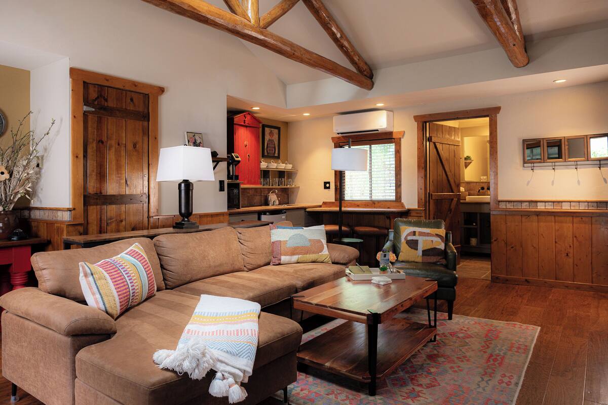 A cozy living room with a brown leather sofa, a wooden coffee table, and a patterned rug. There is a kitchen area visible through an open doorway, featuring wooden cabinets and a red door.