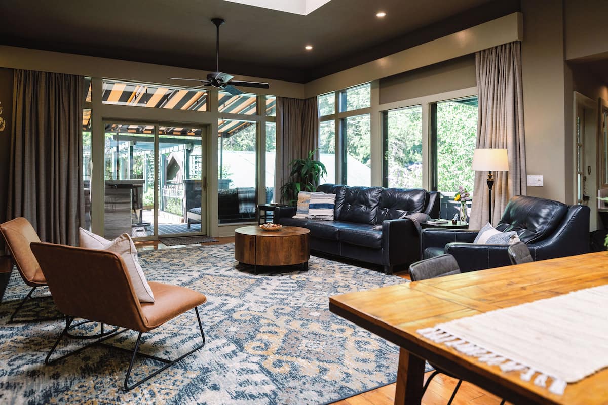 A spacious living room with a large sofa, two armchairs, a wooden coffee table, and a dining table. Large windows provide natural light and a view of the outdoors.