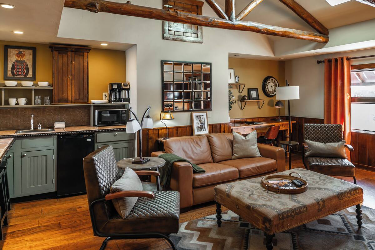A spacious living area with a brown leather sofa, two armchairs, a coffee table, and a dining table. A kitchen with a dishwasher and a microwave is visible in the background.