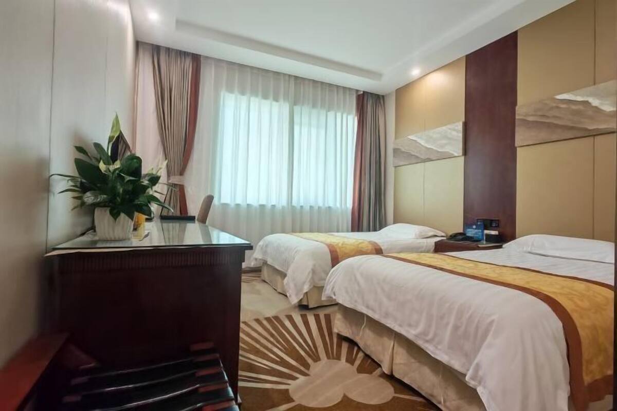 A hotel room with two beds, a desk, a chair, a potted plant, and a large window with curtains.