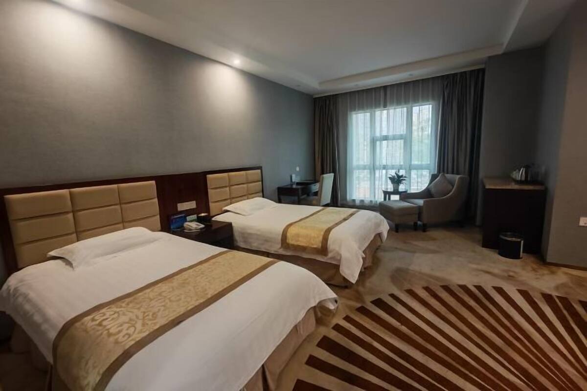 A hotel room with two beds, a desk, a chair, a window with curtains, and a patterned rug.