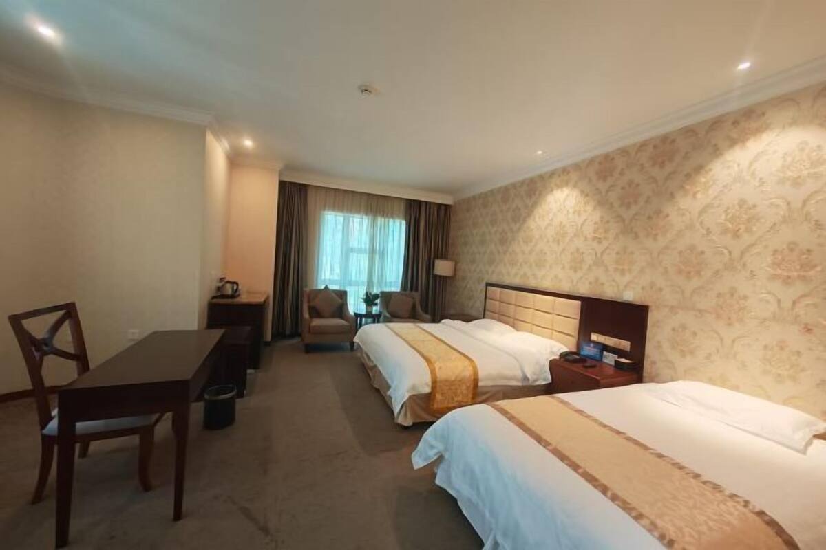 A hotel room with two beds, a desk, a chair, a lamp, and a window with curtains.