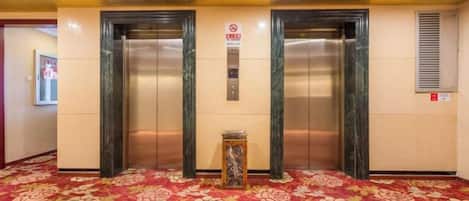 Un espace dédié aux ascenseurs, comprenant deux ascenseurs, un sol recouvert de moquette et un panneau fixé au mur.