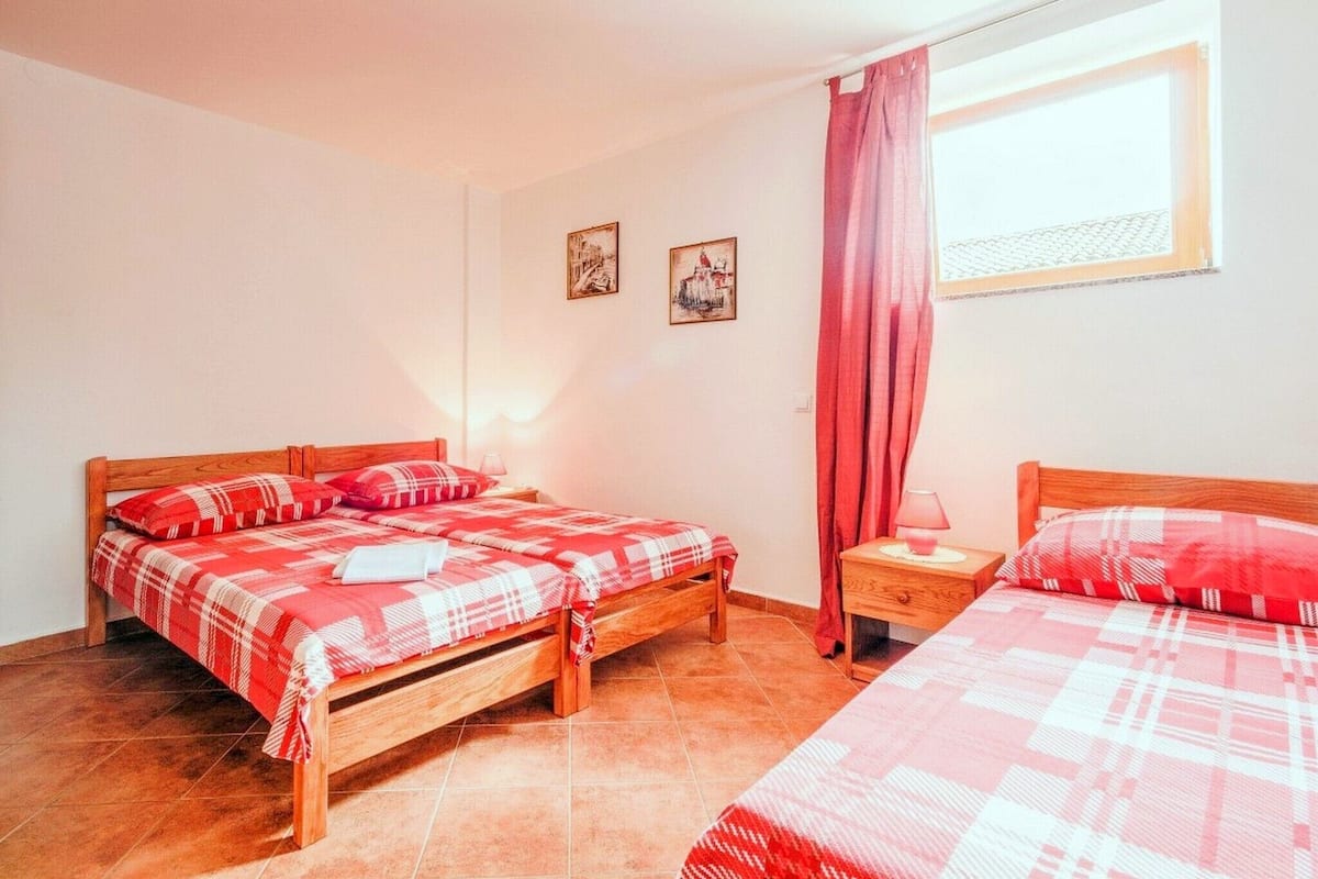 A room with two wooden beds, each with a red and white checkered bedspread and matching pillows. There are two wall-mounted lamps, a window with a curtain, and two framed pictures on the wall.