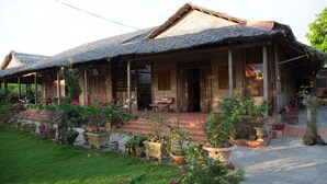 A traditional wooden house with a thatched roof, a porch with potted plants, and a well-maintained garden. - Phuong Thao Homestay (An Binh)