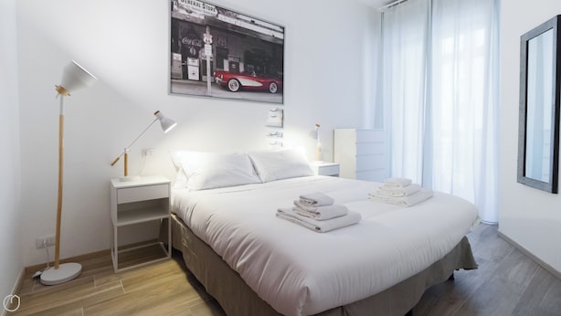 Een moderne hotelkamer met een net opgemaakt bed, een bijzettafeltje met een lamp, een aan de muur hangend schilderij en een raam met gordijnen.