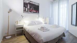 A modern hotel room with a neatly made bed, a side table with a lamp, a wall-mounted picture, and a window with curtains.