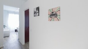 Een gang met een witte muur, een deur en een raam met gordijnen. Twee ingelijste foto's hangen aan de muur.