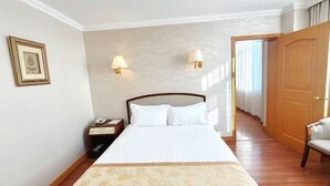 Een hotelkamer met een groot bed, twee bedlampen, een nachtkastje met een telefoon, een lijst met foto op de muur en een houten deur.