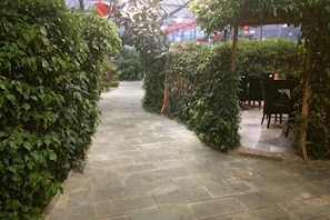 A paved pathway flanked by dense green hedges, leading to a covered outdoor seating area with tables and chairs. - Xinshuangquan Hostel (Beijing)