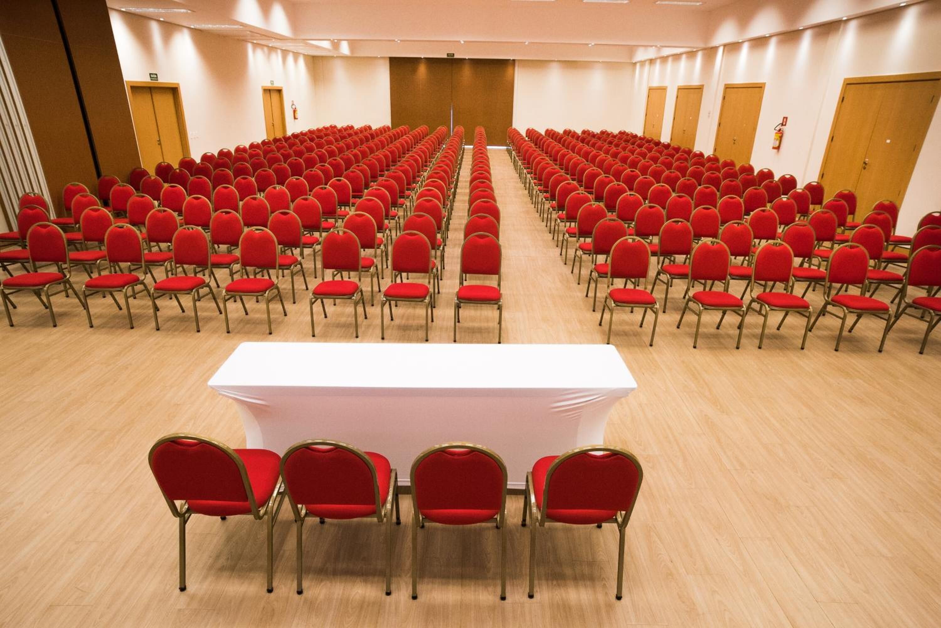 Un salón de conferencias con filas de sillas rojas y una mesa blanca en el centro.