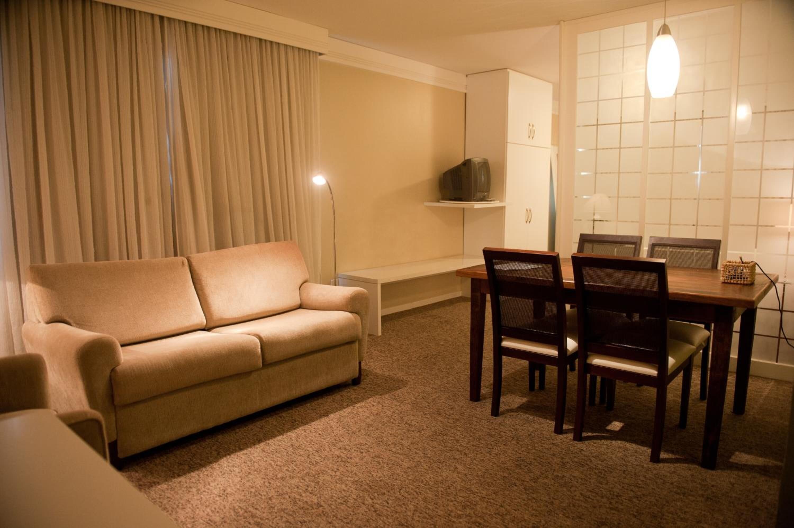 Una sala de estar con un sofá color crema, una mesa de comedor con cuatro sillas, un mueble pequeño y un televisor sobre una estantería.