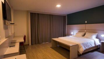 A modern hotel room with a large bed, bedside tables, a desk, a chair, a TV, and a window with curtains.