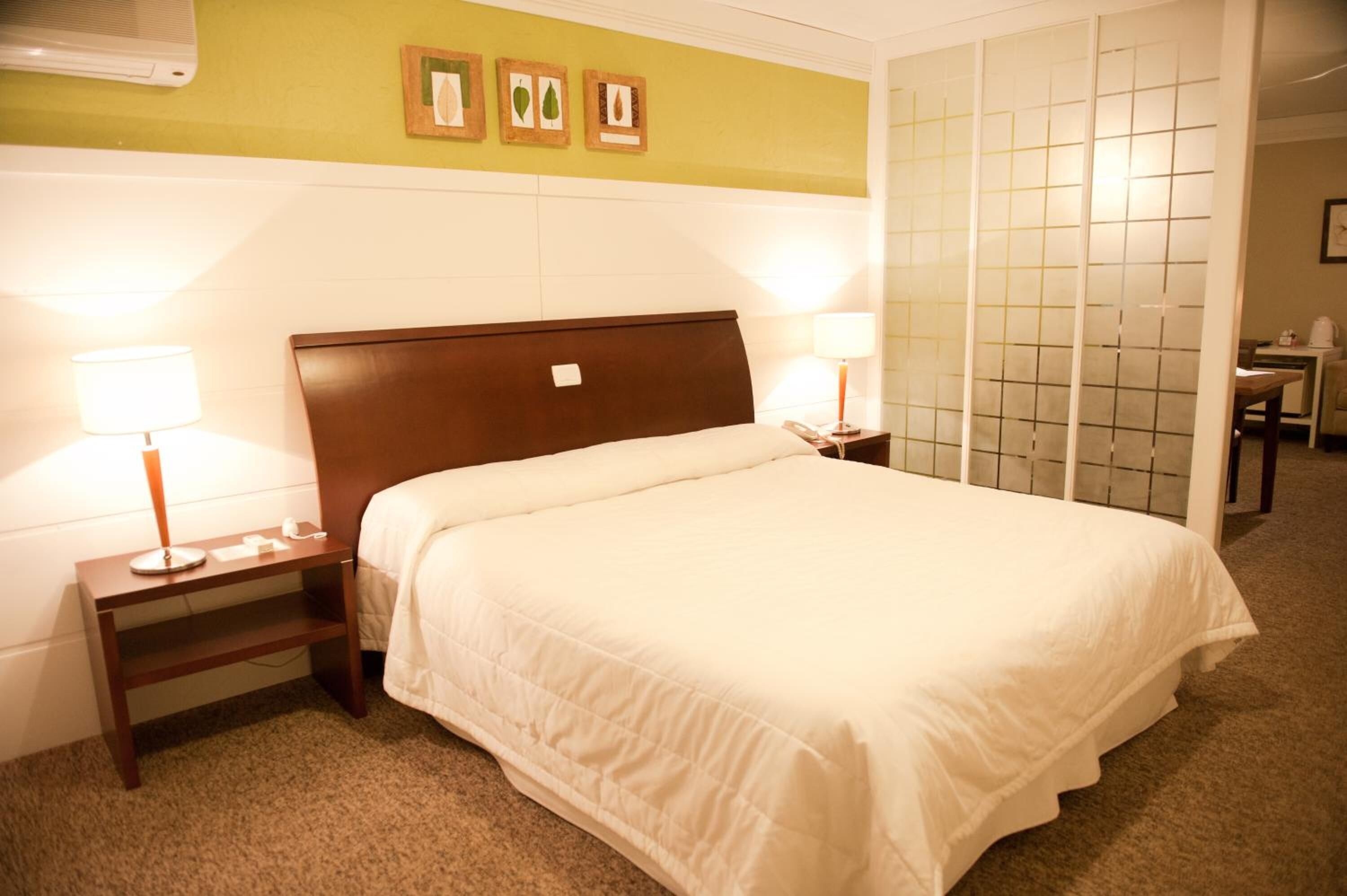 Quarto de hotel com uma cama grande, duas mesas de cabeceira com luminárias, uma pequena escrivaninha e uma cadeira.