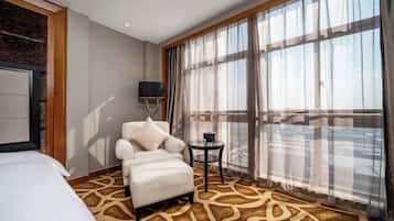 A hotel room with a bed, a white armchair, a small table, a TV, and large windows with sheer curtains.