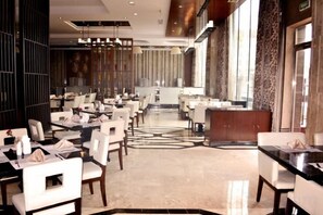 Un vaste espace de restauration dans l’hôtel, avec de nombreuses tables et chaises, une allée centrale et de grandes fenêtres laissant entrer la lumière naturelle.